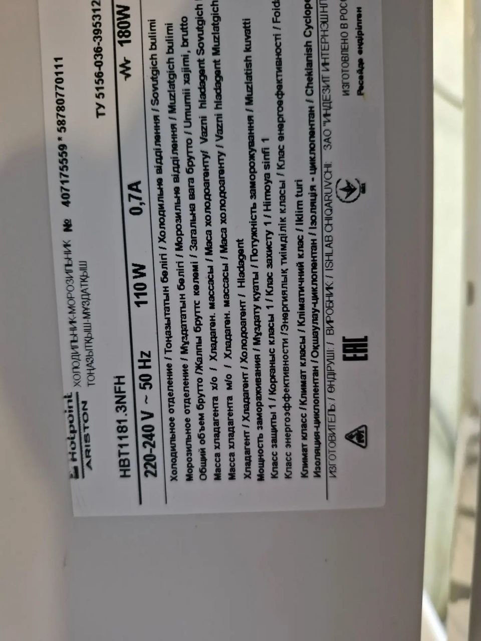 Дверца холодильника с логотипом Hotpoint Ariston и шильдиком модели; открытая нижняя панель и область с компрессором во время ремонта, ракурс 2