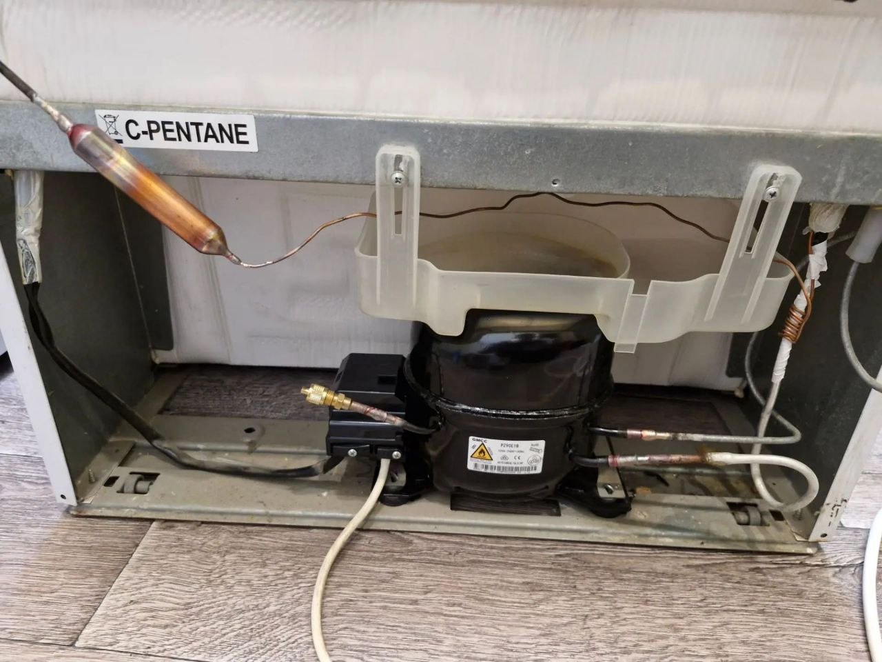 Дверца холодильника с логотипом Hotpoint Ariston и шильдиком модели; открытая нижняя панель и область с компрессором во время ремонта, ракурс 3